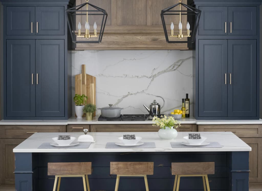 Navy blue shaker cabinets flanking a white marble-look backsplash, with a navy island, warm wood stools and brass lantern pendants