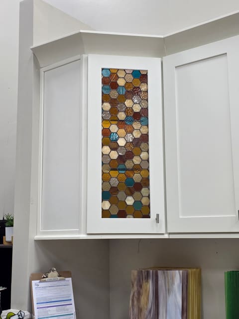 White shaker corner cabinet with a stained glass door insert of amber, rust, teal and cream honeycomb hexagons